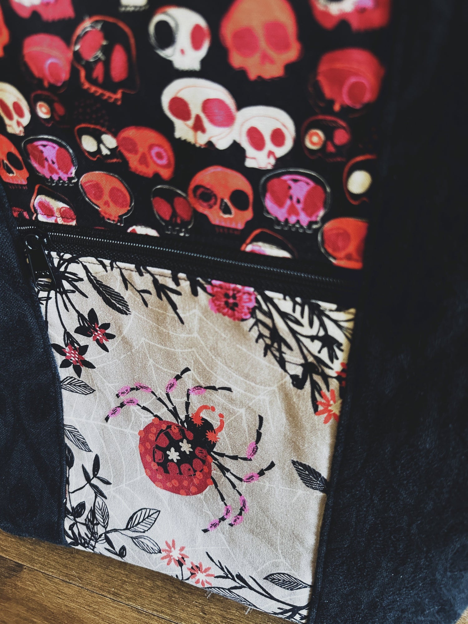 Image of the front of a halloween tote bag. It features spider fabric on a zipper pocket and skull accent fabric in deep reds, oranges, pinks, and blacks on an ivory background. 