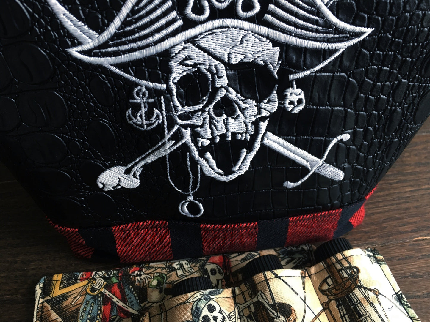 Image of a large handmade pirate themed essential oil organizer. It features a large skull and crossbones embroidery on faux alligator leather, bold red and black skull woven textile. The inside is made from blood red linen and skelewags fabric from Alexander Henry. It  has 10 built in pockets and 3 inserts with 3 pockets each.  