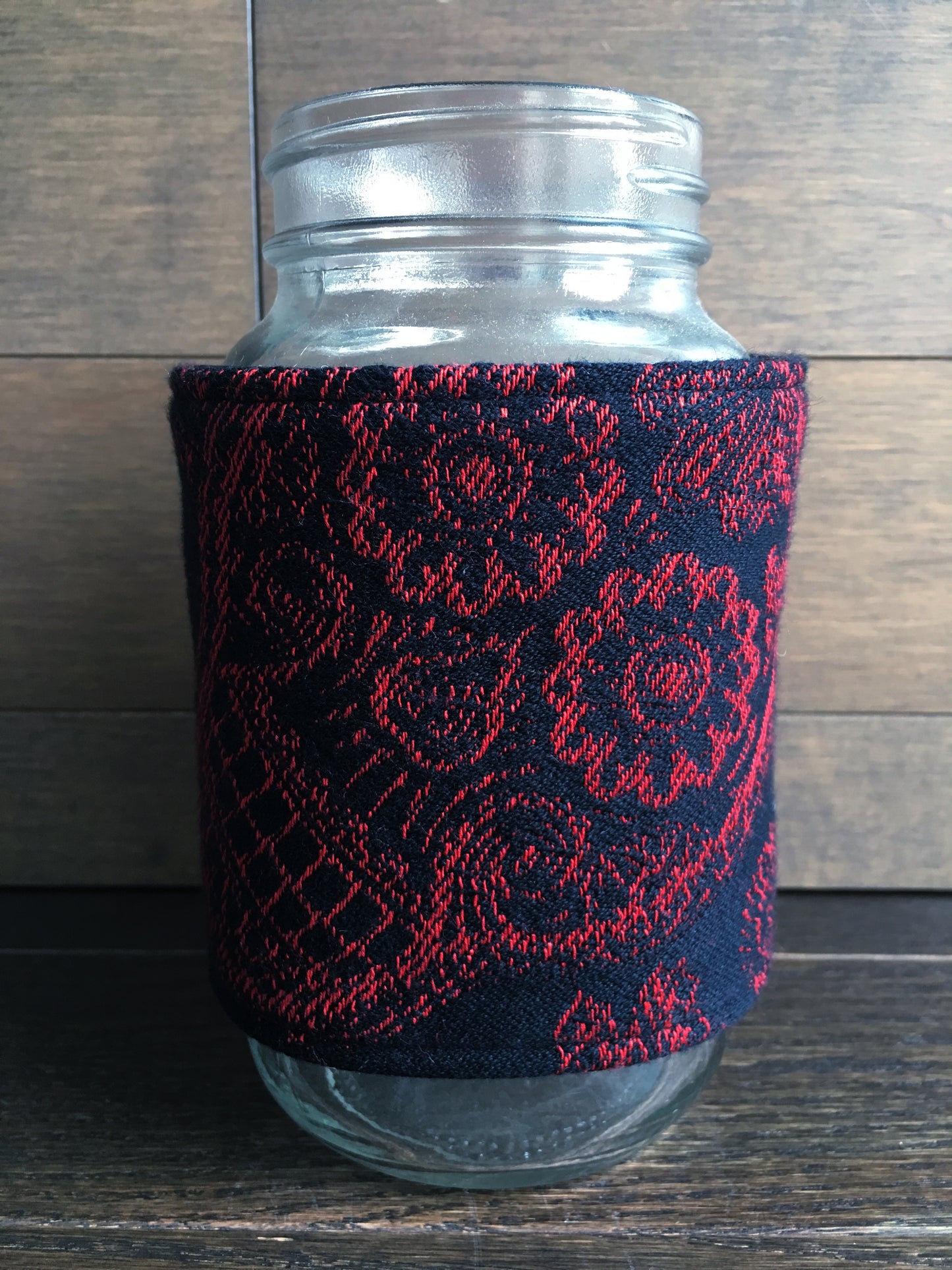 Image of a handmade and pirate themed mug or mason jar cozy. It features bold red and black skull woven jacquard on one side and a red skull and crossbones pirate crest embroidered on black faux alligator leather on the reverse. 