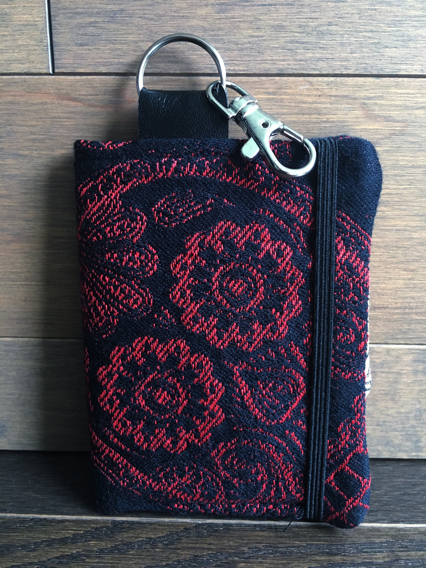 Image of a handmade and pirate themed mini card wallet made from red and black jolly roger woven textile, skelewag fabric, and black faux leather. It features a machine embroidered skull and crossbones and a wallet clip. 