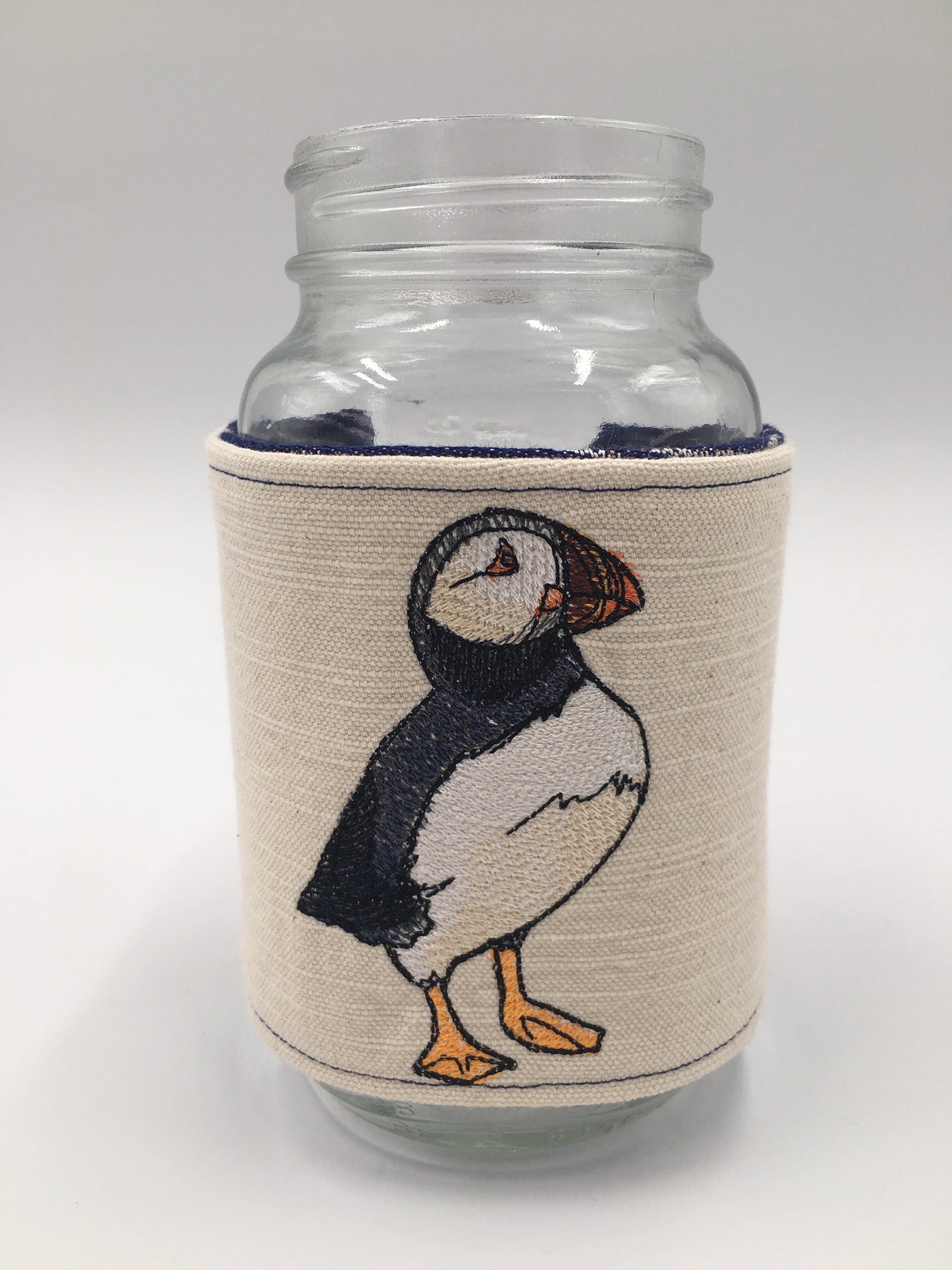 Image of a handmade jar and mug cozy featuring oscha slings puffins woven fabric in deep blue, ivory, and orange and elegant and realistic puffin embroidery. 