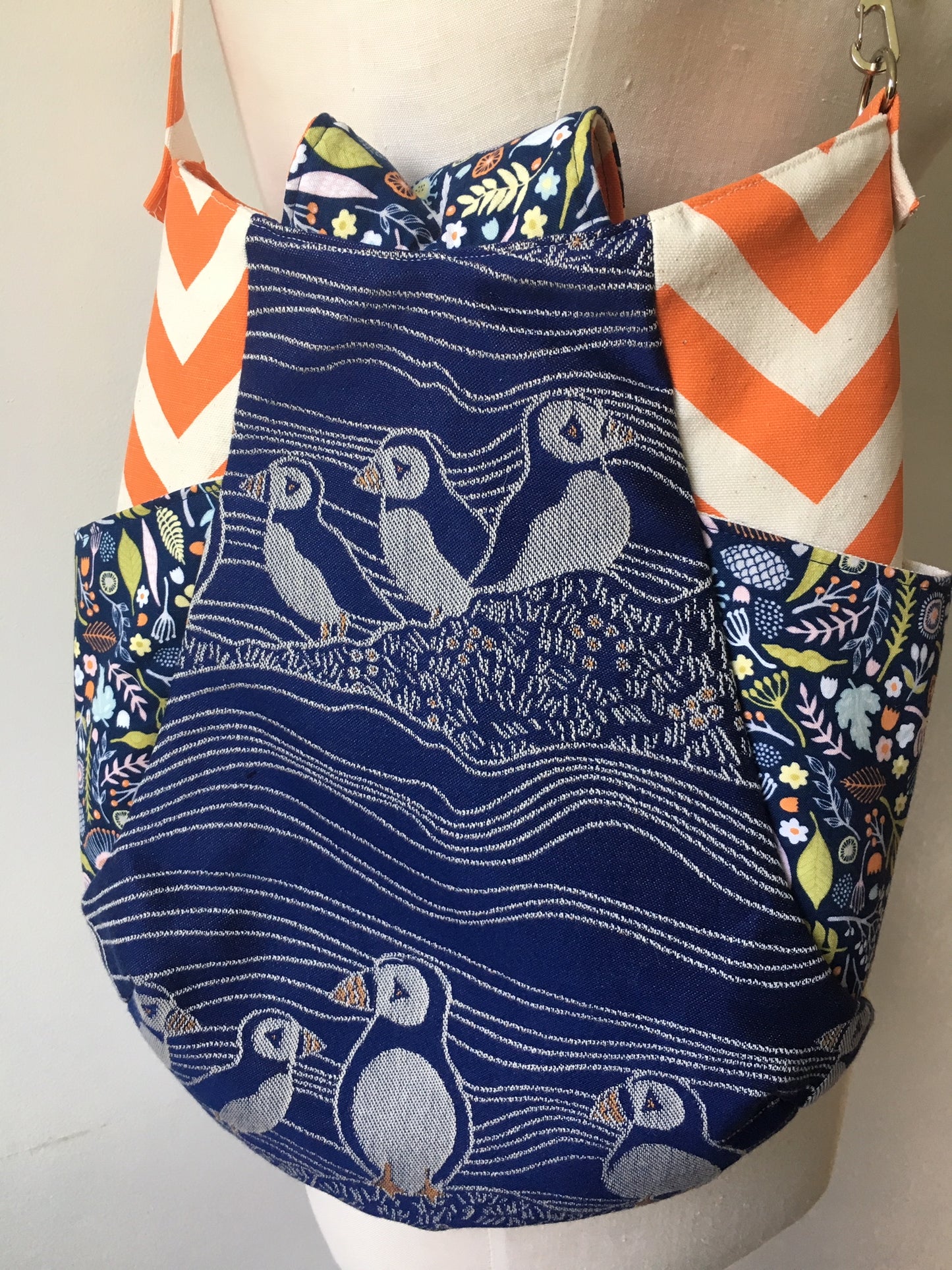 Image of a beautiful handmade boho hobo style crossbody bag featuring puffin wrap scrap from Oscha Slings and elegant puffin machine embroidery. It also has vibrant orange side panels, two side slip pockets, and a fold over flap top closure. 