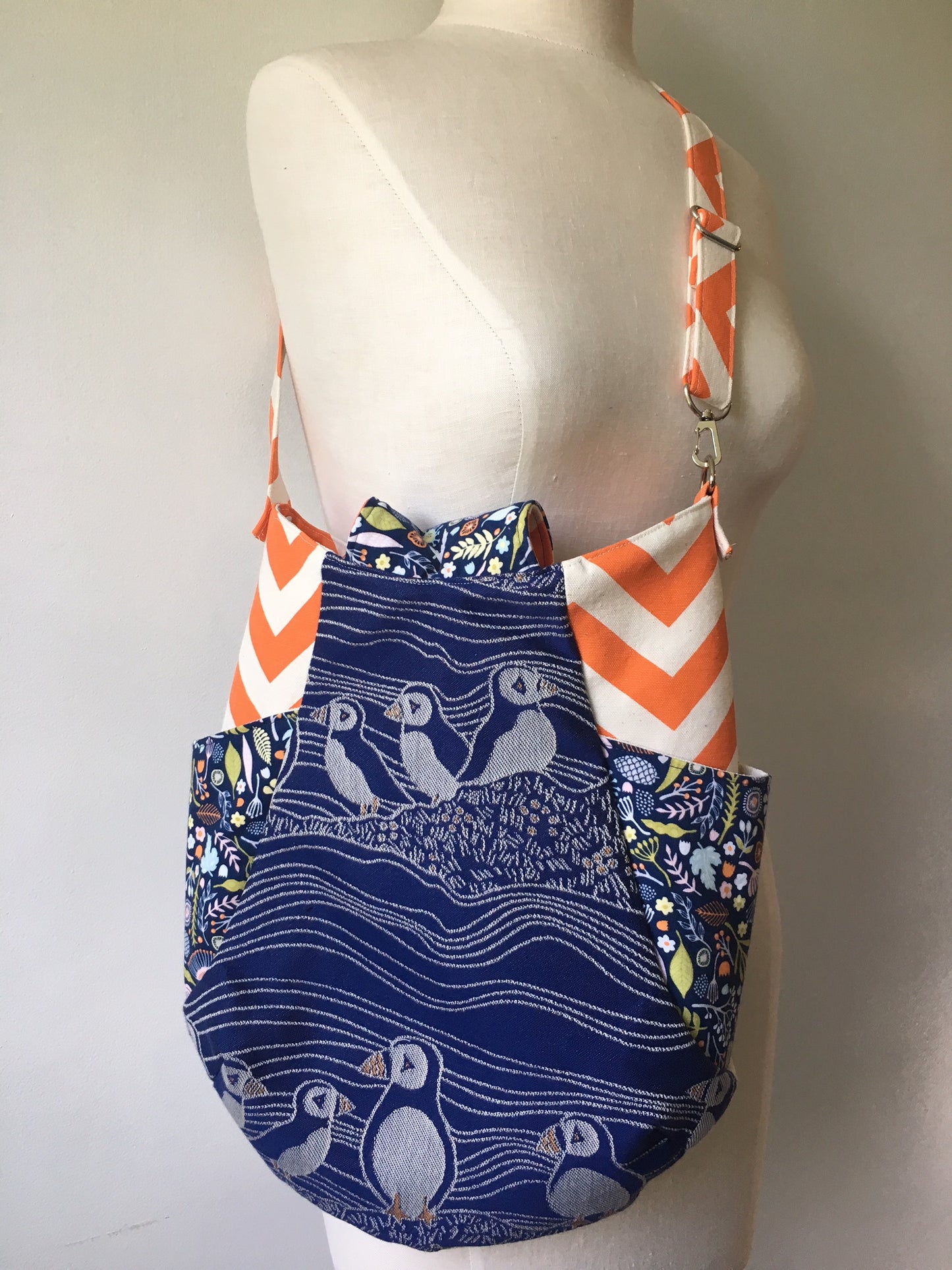 Image of a beautiful handmade boho hobo style crossbody bag featuring puffin wrap scrap from Oscha Slings and elegant puffin machine embroidery. It also has vibrant orange side panels, two side slip pockets, and a fold over flap top closure. 